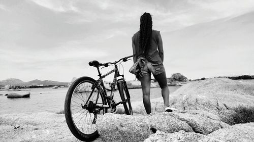 Rear view of young woman standing by bicycle on rock formation against sky