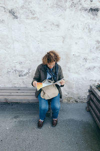 High angle view of woman reading map while sitting on bench against wall