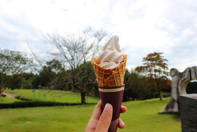 Cropped hand of woman holding ice cream cone against cloudy sky at park