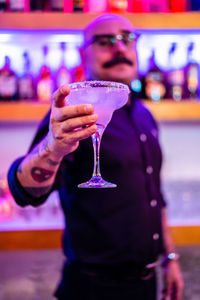 Confident stylish barkeeper with glass of cold alcoholic cocktail standing against shelves with various drinks