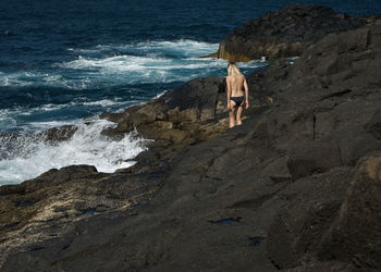 Rear view of shirtless woman walking on rocks by sea