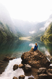 Man sitting on rock by lake against mountain