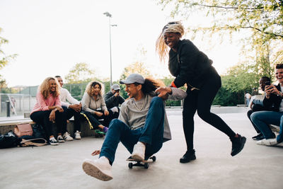 Teenage girl pushing young man on skateboard while friends laughing at park