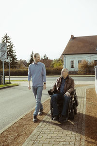 Smiling senior man with disability in motorized wheelchair by young caregiver walking on road during sunny day