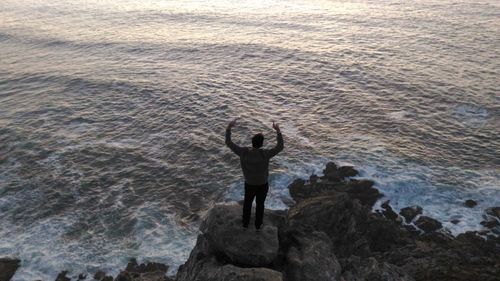 High angle view of man with arms raised standing at rocky beach
