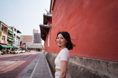 Side view young cheerful asian female millennial with dark hair in casual clothes smiling happily and looking at camera against red background