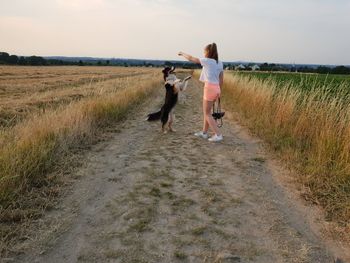 Girl playing with dog on dirt road against sky during sunset
