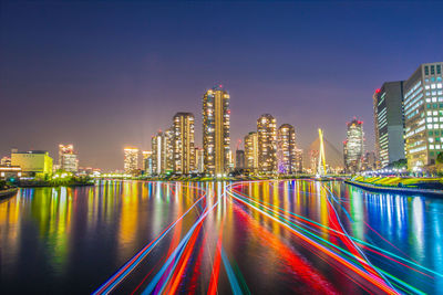 Light trails over river by illuminated buildings at night