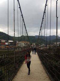 People walking on footbridge against suspension bridge