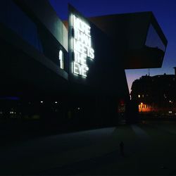 Illuminated road by building against sky at night