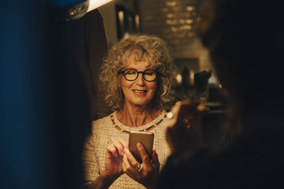 Smiling senior woman using smart phone while sitting at illuminated home during party