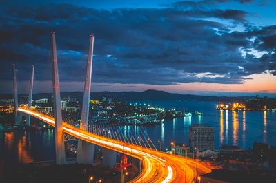 Light trails on bridge in city against cloudy sky at night