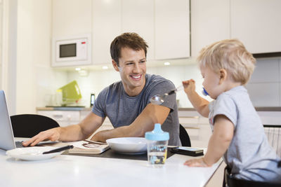 Happy father looking at baby boy while using laptop in kitchen