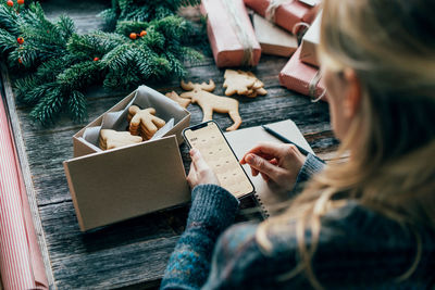 Woman writing plans for 2024 on the christmas table, holding a phone with a calendar on the screen.