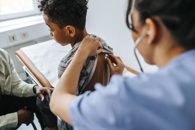 Female pediatrician examining boy's back with stethoscope in examination room