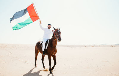 Man riding horse on sand at beach against clear sky