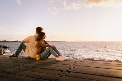 Couple sitting back to back on seat while looking at sea during sunset