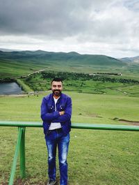 Full length portrait of smiling man standing on grassy field against cloudy sky