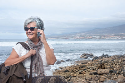 Senior woman talking over smart phone at beach against sky