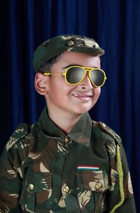 Smiling boy in army uniform and sunglasses standing against curtain