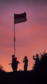 Silhouette children standing on hill by flag against sky during sunset