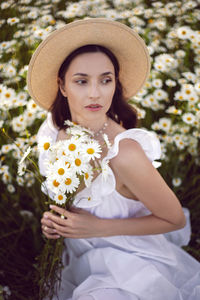 Beautiful young brunette woman in a hat and a white dress standing on a chamomile field at sunset