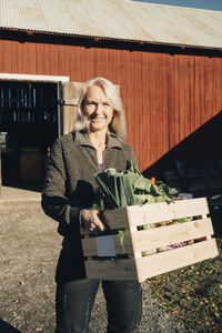 Portrait of smiling mature woman carrying crate full of vegetables with barn in background