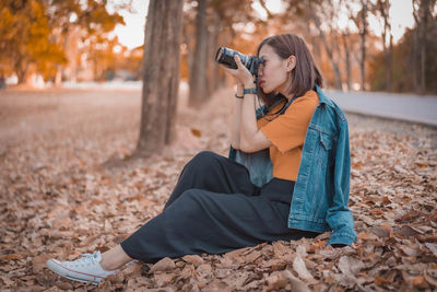 Full length of woman photographing while sitting on dry leaves