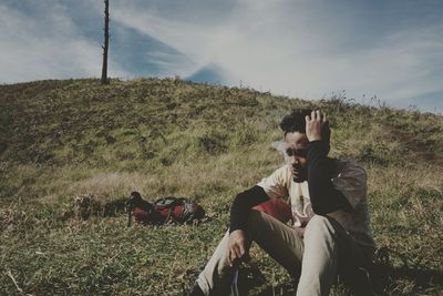 Man smoking while sitting on grassy field against sky at park