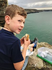 Boy having sandwich while sitting on cliff by sea