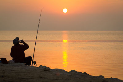 Rear view of silhouette man fishing in sea against sunset sky