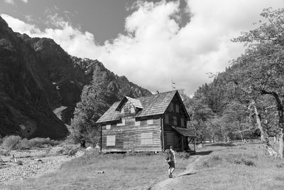 Rear view of hiker walking towards house by mountains against cloudy sky