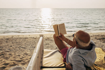 Relaxed senior man lying on bed while reading book on beach at sunset