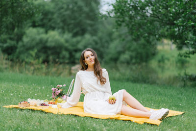 A young beautiful woman in a white dress sits on a blanket on a summer picnic on the green grass.