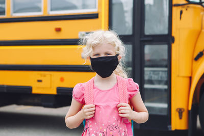 Happy girl student in cloth face mask by yellow school bus outdoors. 