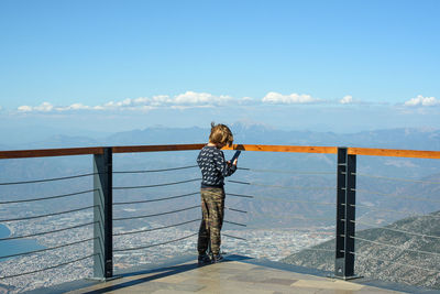 Rear view of boy standing by railing against sky