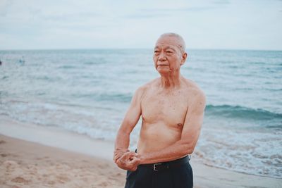 Shirtless senior man showing muscles while standing at beach against sky during sunset