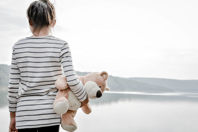 Rear view of girl holding teddy bear while standing against river