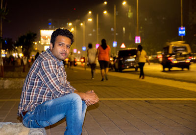 Side view portrait of young man sitting on sidewalk in city at night