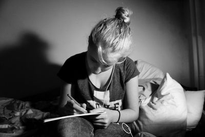 Young woman writing in book while sitting on bed at darkroom