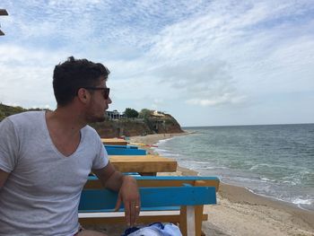 Man sitting on bench at beach against sky