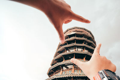 Low angle view of hand gesturing by building against sky