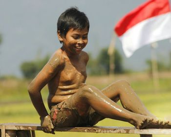 Full length of shirtless boy sitting with indonesian flag in background