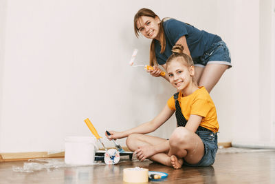 A girl with her sister in bright blue and yellow clothes helps to paint the walls in her room white.