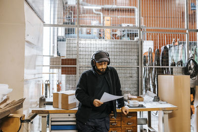 Male employee wearing wireless headphones and cap while reading document at distribution warehouse