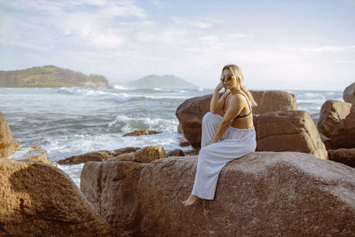 Side view of woman sitting on rock by sea against sky