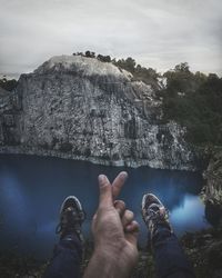 Low section of man gesturing on cliff by lake against cloudy sky