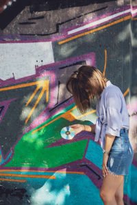 Beautiful woman holding compact disc while standing against graffiti wall