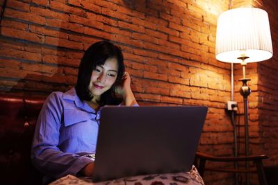 Young woman using phone while standing against brick wall