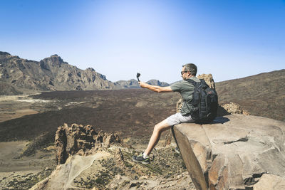 Man taking selfie with camera while sitting on cliff against mountains